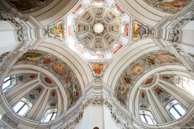 Upward view of an octagonal cathedral dome surrounded by white stucco arches and colorful painted vault panels.