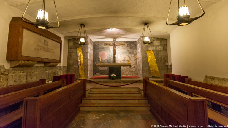 Small vaulted chapel with wooden benches facing a raised altar and crucifix against rough stonework.