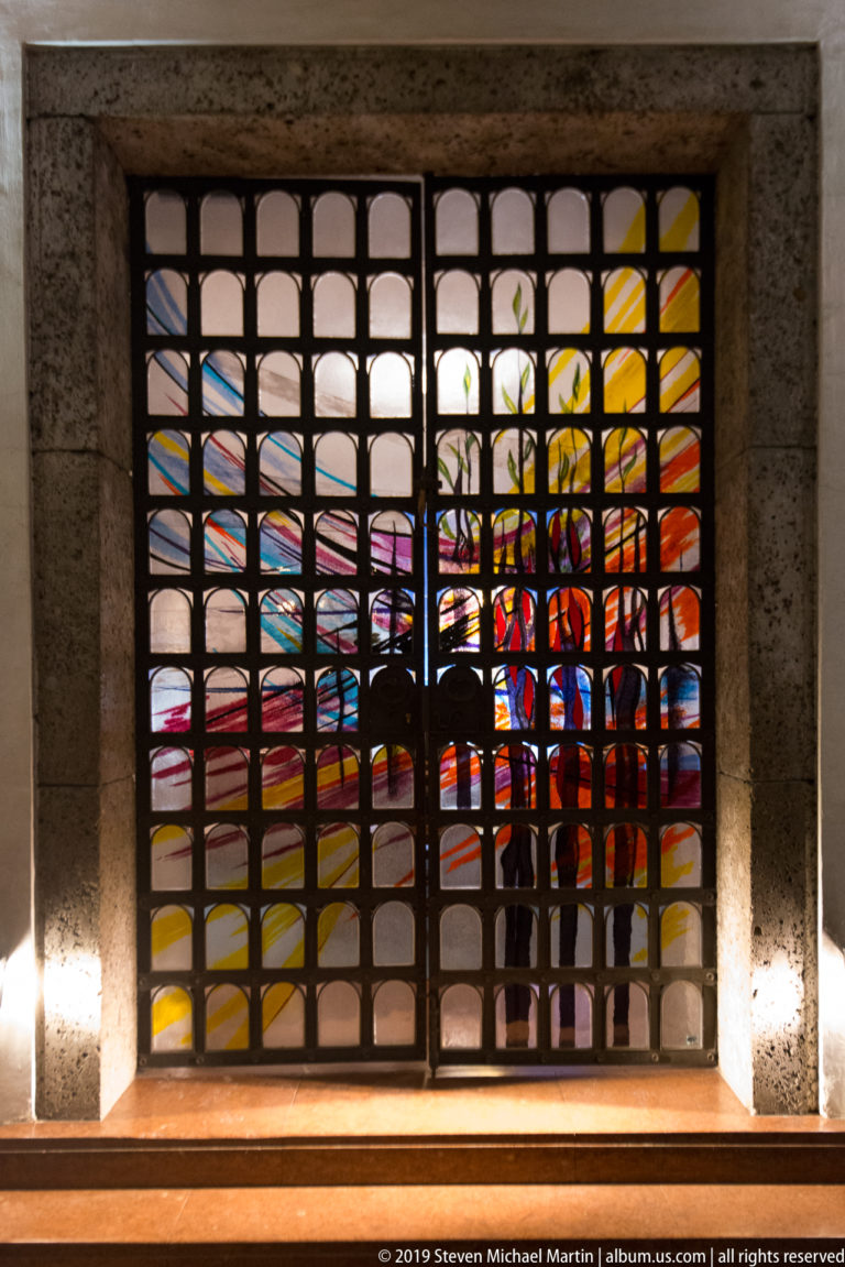 Square black metal grille in front of a brightly colored stained-glass window with red, yellow, blue, and white glass.
