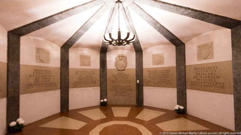 Small polygonal memorial chamber with Latin inscriptions, heraldic panels, dark ribs, and a central hanging light.