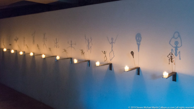 Diagonal row of small wall lights beneath elongated line-drawn figures on a dim blue-gray wall.
