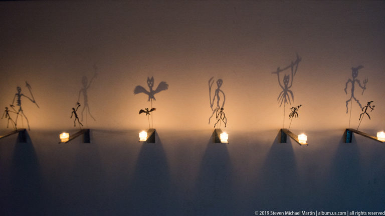 Small wall lights illuminate thin drawn human and animal figures across a dark interior wall.