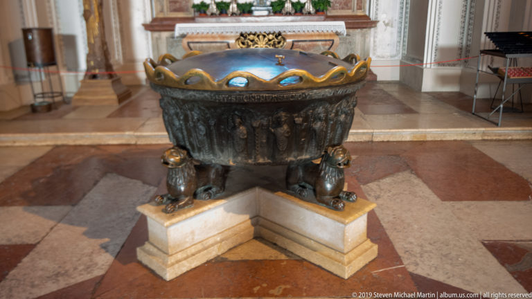 Dark bronze baptismal font on four lion supports, raised on a pale stepped base inside Salzburg Cathedral.