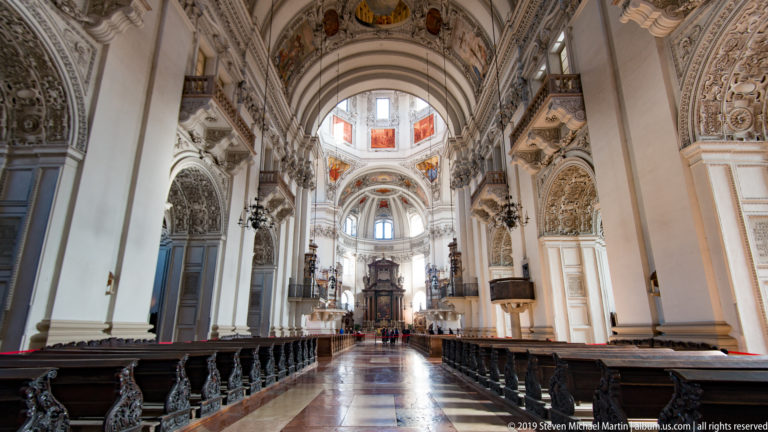 Central aisle of Salzburg Cathedral with wooden pews leading toward the high altar and bright crossing dome.