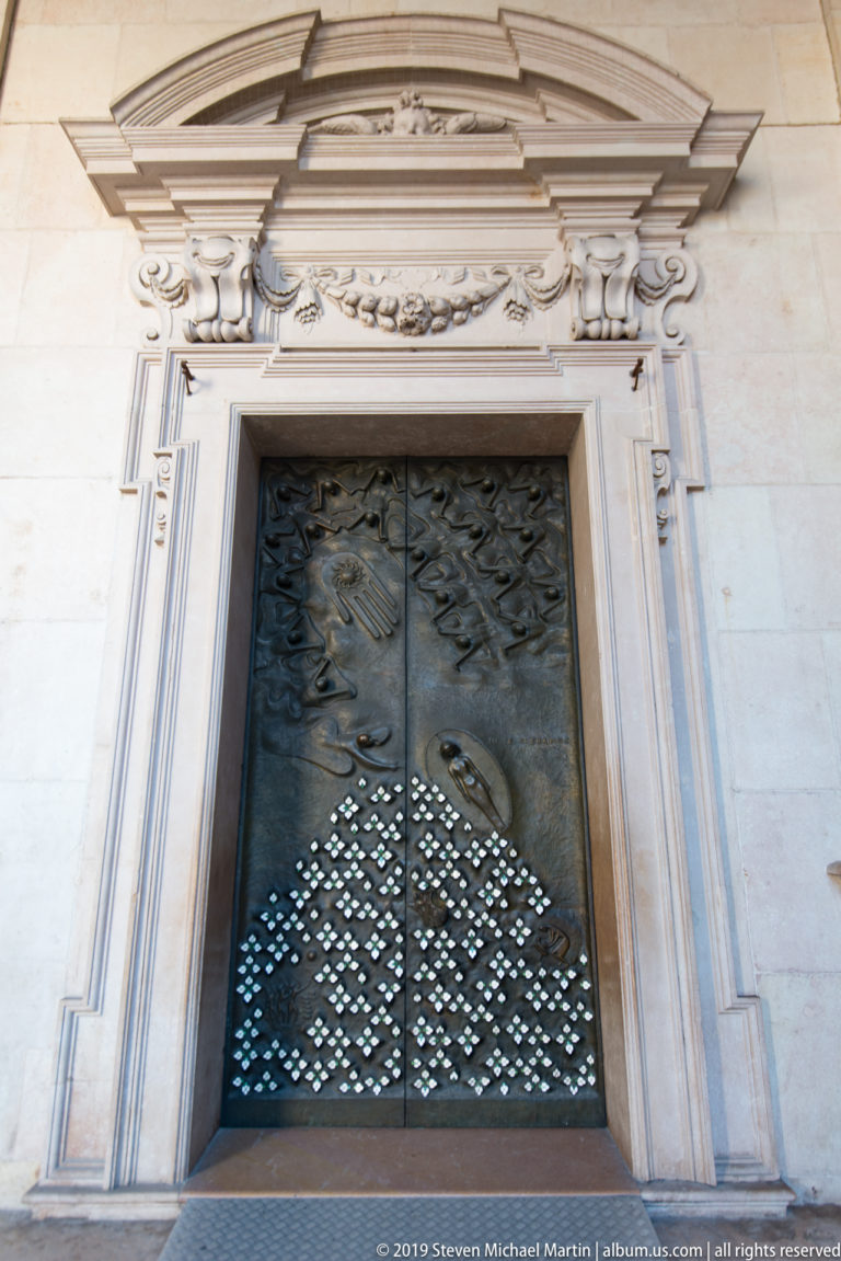 Dark metal double doors with abstract relief and perforated lower panels framed by an ornate pale stone portal.