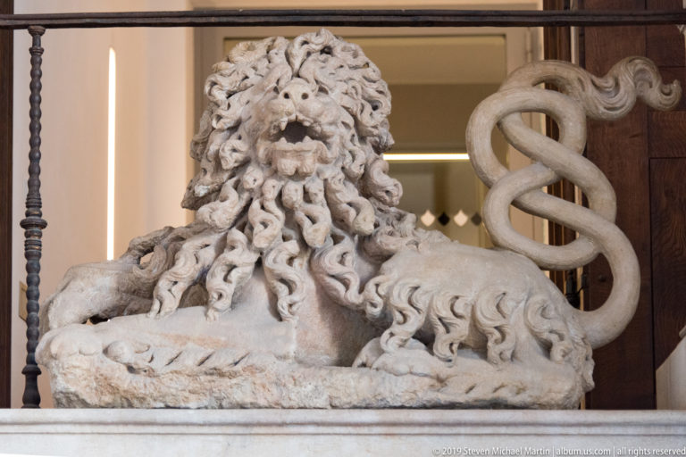 Carved stone lion lying down with a dense curled mane and looping tail beside its body.