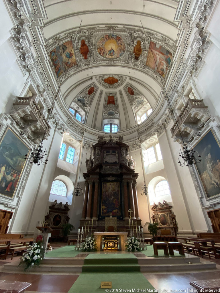 Closer frontal view of high altar with dark architectural frame, painted altarpiece, flowers, bright arched windows, chandeliers, and side furnishings.