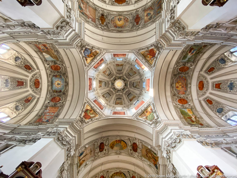 Straight-up view of octagonal dome center with four radiating decorated vault arms, painted scenes, medallions, ribs, and white stucco.