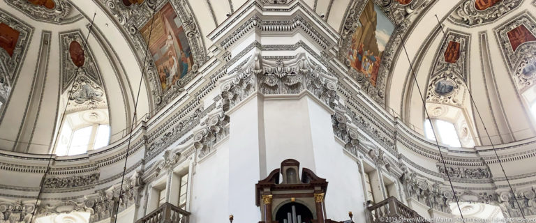 Steep upward view of a massive crossing pier with layered stucco cornices, painted vaults, tall windows, and dark organ case below.