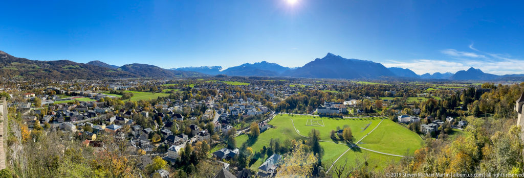 Wide sunny panorama from Fortress Hohensalzburg over Salzburg neighborhoods and green fields toward jagged Alpine peaks.