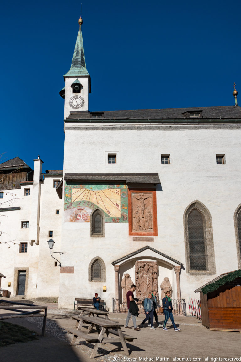 Visitors walk before a white fortress façade with clock tower, mural, relief panels, pointed windows, benches, and wooden stall.
