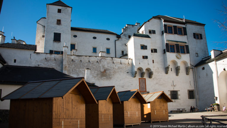 Wooden market stalls stand in a fortress courtyard below tall whitewashed buildings with projecting bays and a square tower.