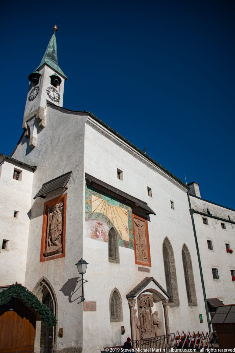 White fortress courtyard façade with clock tower, tall pointed windows, painted panels, and sculpted entrance decoration.