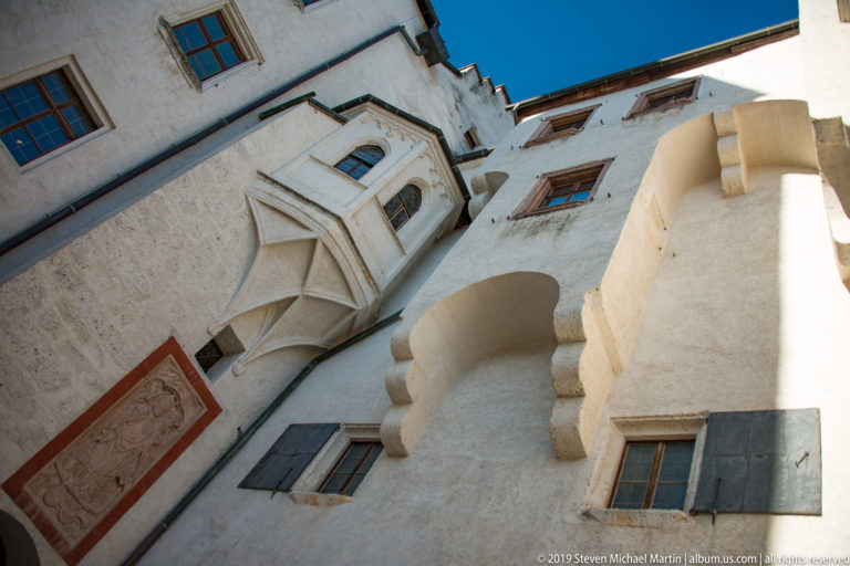 Upward view of a projecting oriel window on a white fortress wall, with stepped corbel, shutters, and painted panel.