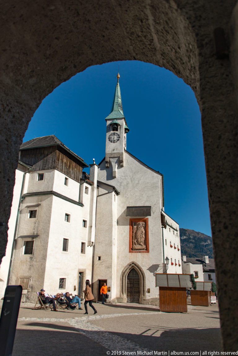 White chapel with clock tower and relief panel seen through a stone arch inside Hohensalzburg Fortress.