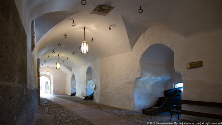 Long white vaulted corridor with hanging lanterns, ceiling rings, deep wall recesses, benches, and an artillery display.