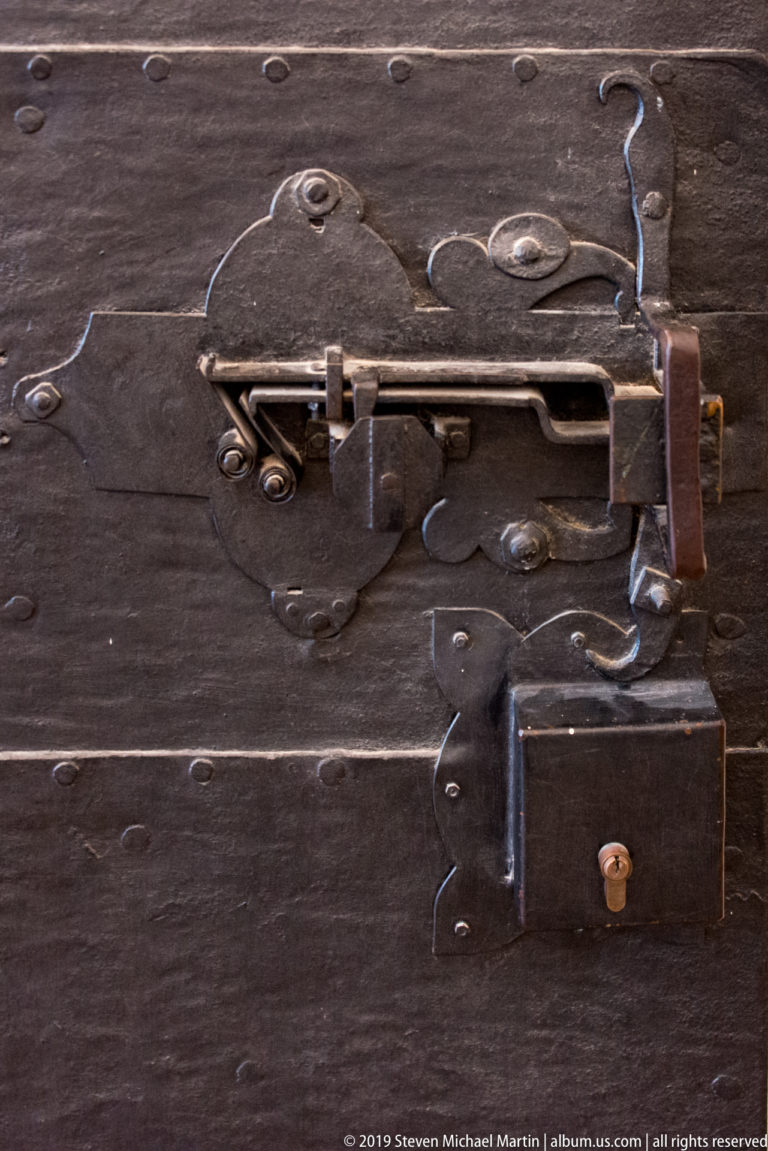Close view of heavy iron lock, latch, straps, and fasteners mounted across a dark wooden fortress door.