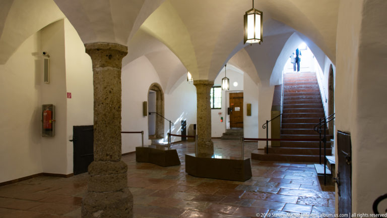 Stone column beneath white vaults in a fortress hall, with lanterns, display cases, and stairs rising at right.
