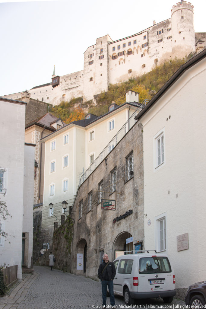 Fortress Hohensalzburg stretches across the hilltop above a paved court, lower buildings, parked vehicle, and wooded slope.