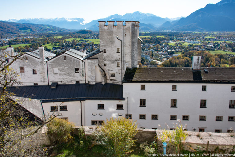 Clustered white fortress buildings and a tall square tower seen from above, with Salzburg’s valley and mountains beyond.
