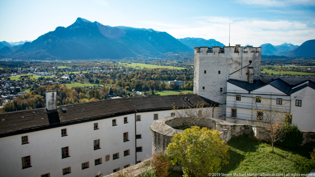 White fortress buildings and a square tower overlook the Salzburg basin with mountain ridges in the distance.