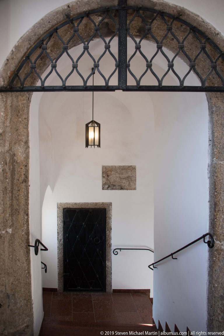Steps descend beneath a stone arch and iron grille toward a dark doorway inside Fortress Hohensalzburg.