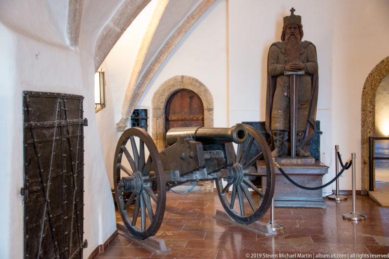 Historic wheeled cannon displayed in a vaulted fortress room beside a large crowned figure holding a sword.
