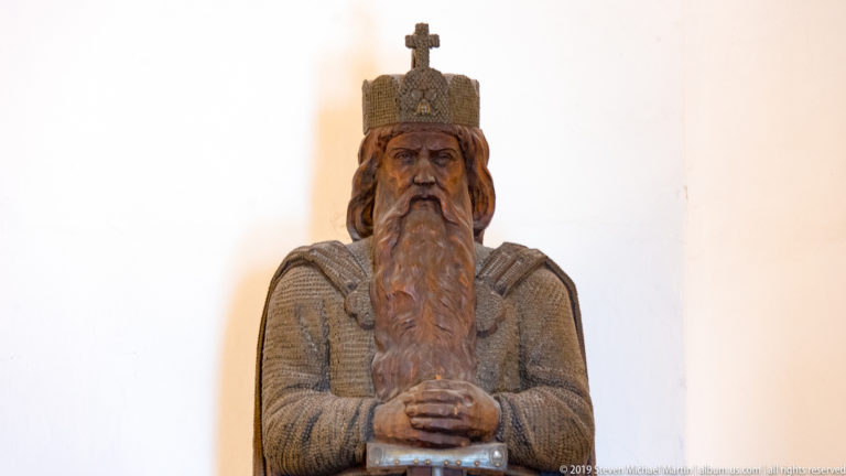 Carved crowned and bearded male figure standing frontally against a white museum wall at Fortress Hohensalzburg.