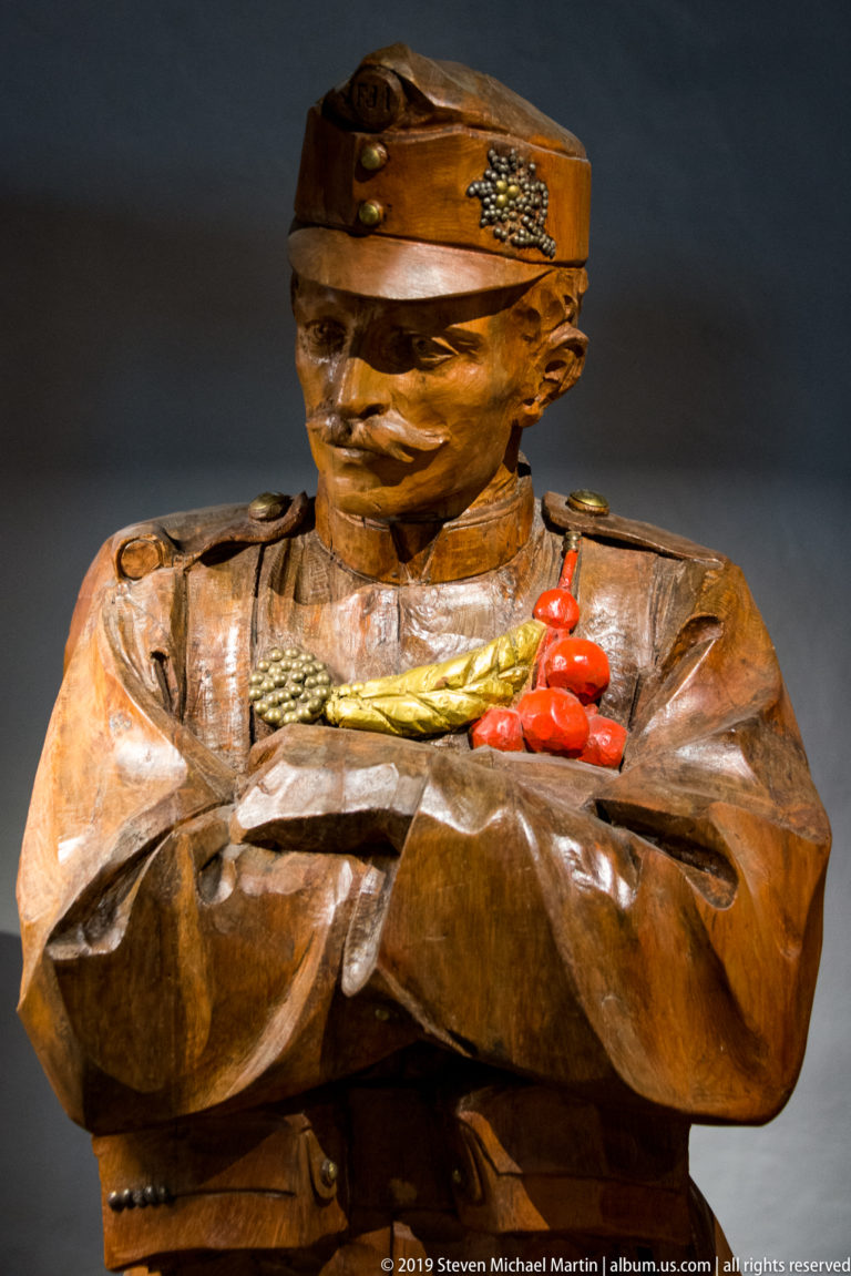 Carved wooden bust-length soldier in uniform and cap, arms crossed over a cluster of bright red objects.