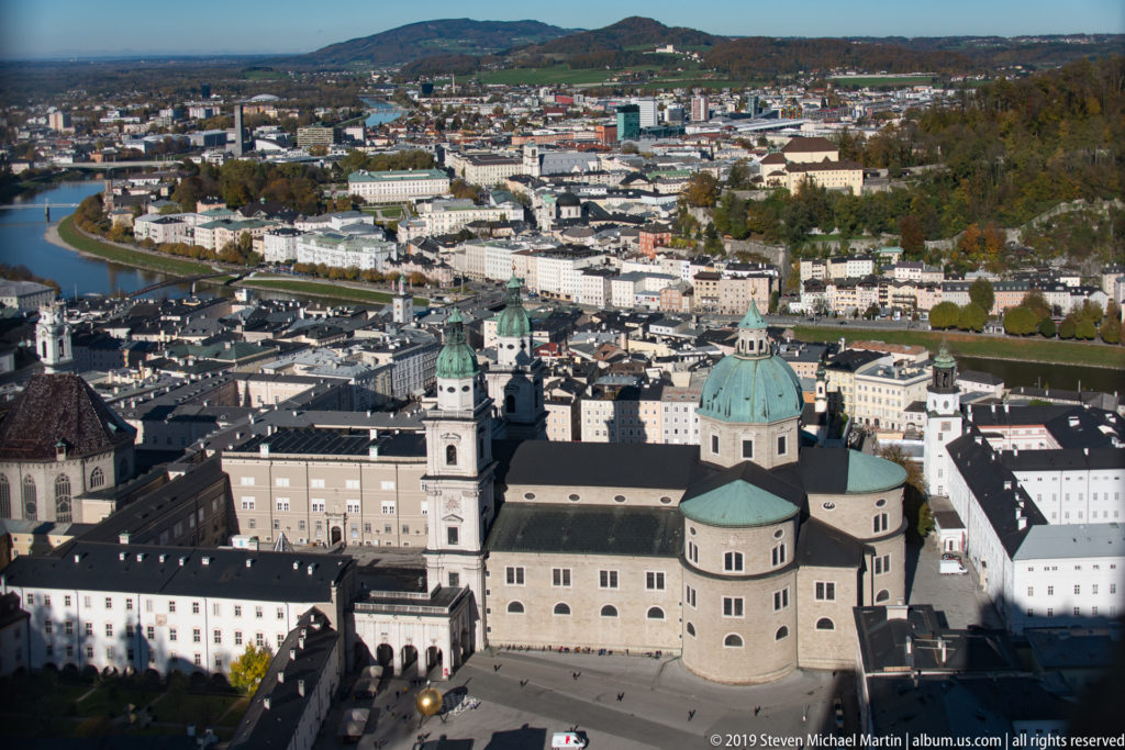 Elevated view from Hohensalzburg Fortress over Salzburg Cathedral, old-city roofs, the Salzach, and the modern city beyond.