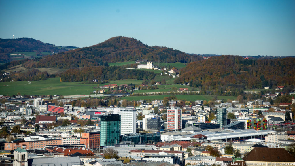 Elevated panorama of modern Salzburg neighborhoods, green fields, and wooded hills beyond the city.