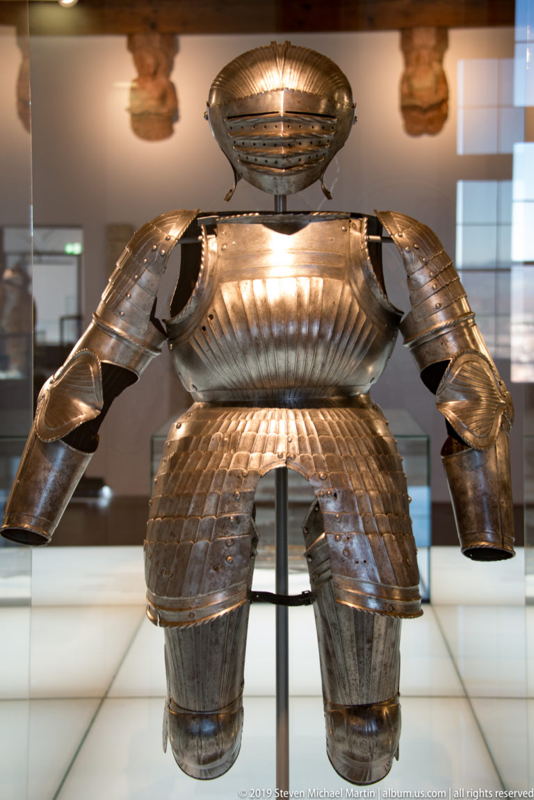 Front view of a polished steel suit of plate armor displayed on a museum stand.