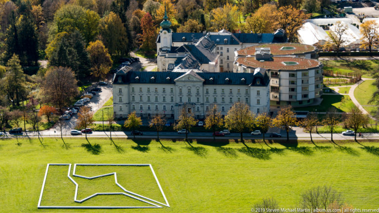 Large pale building with green-roofed tower below the fortress, beside a field marked by a white geometric outline.