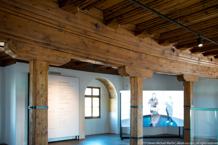 Heavy exposed timber posts and beams inside a modern museum gallery with windows and glass barriers.