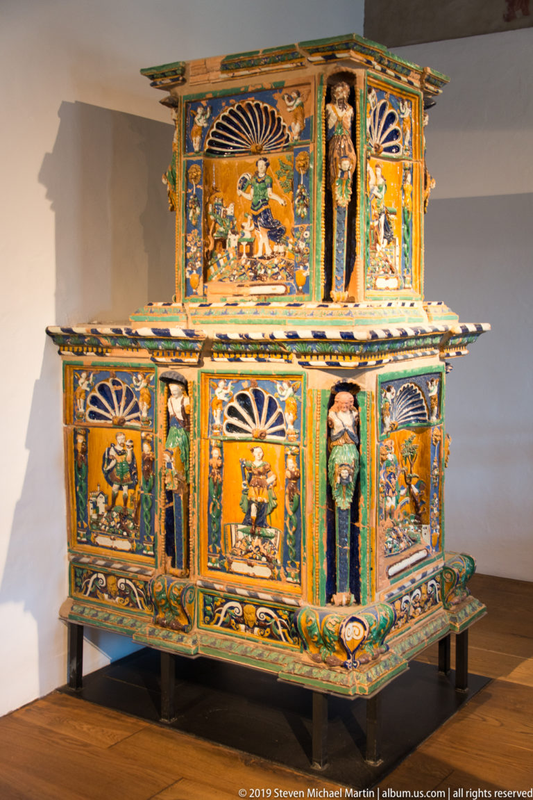 Large two-tiered ceramic tile stove with colorful glazed figures, architectural panels, and floral ornament.