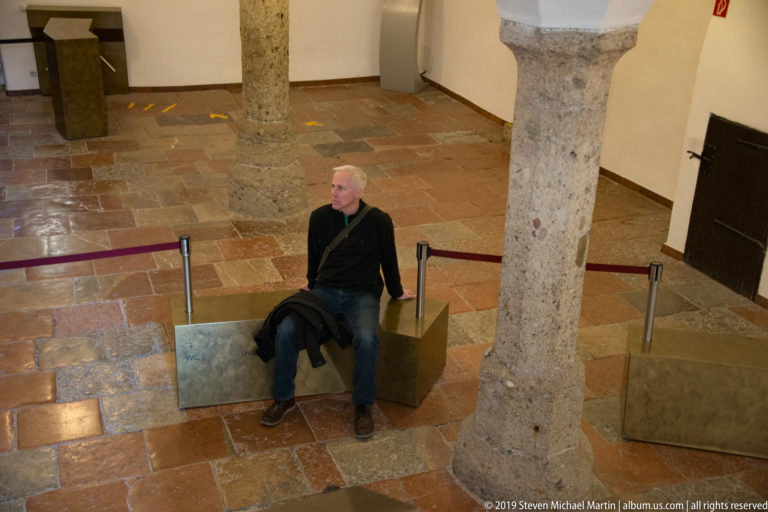 Seated visitor on a bench between thick stone columns in a white-walled room with terracotta-colored floor tiles.
