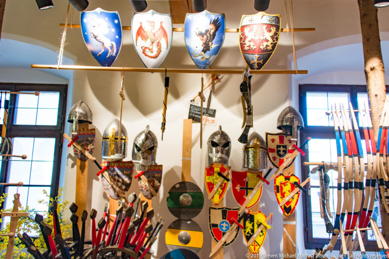 Museum wall filled with helmets, shields, polearms, blades, and colorful heraldic escutcheons.