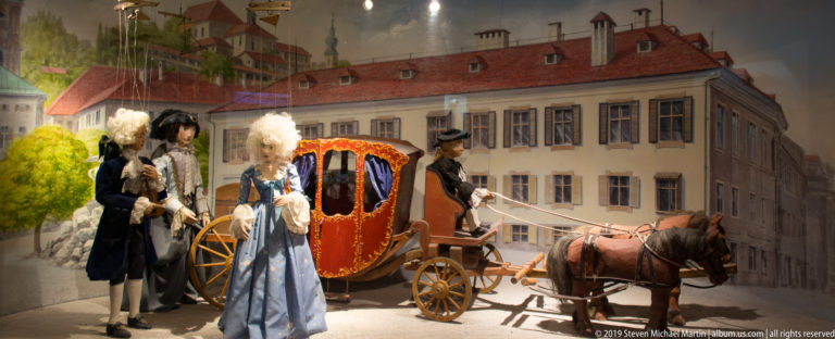 Red-and-gold horse-drawn carriage with costumed figures in powdered wigs before a palace backdrop.