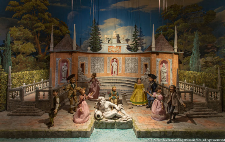 Miniature courtly garden diorama with costumed figures, fountain, terraces, and painted landscape backdrop.
