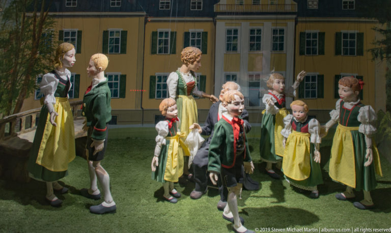 Costumed adult and child puppet figures posed together before the façade of a large building.