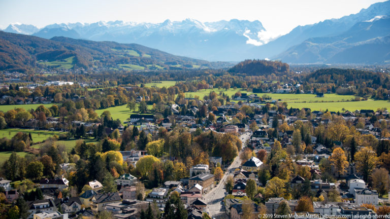 Wide elevated view over Salzburg neighborhoods, green fields, autumn woodland, and distant mountain ridges.