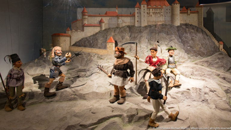 Armed puppet figures on a rocky stage with a model hilltop fortress and mounted figures behind them.