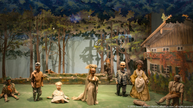 Articulated figures arranged in a rural woodland diorama beside a rustic house in a museum display.