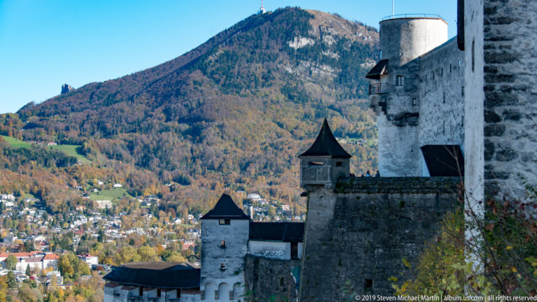 Pale fortress towers and stone walls descending a slope with a wooded mountain rising behind them.