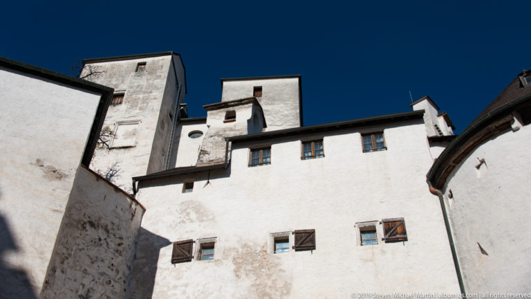 Upward view of weathered white fortress walls and tower-like masses with small shuttered windows under a deep blue sky.