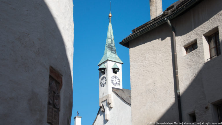 White clock tower with green pointed roof and clock faces framed tightly between fortress buildings against blue sky.