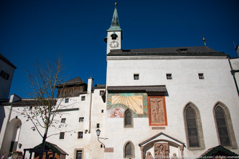 Leafless tree before a white fortress façade with clock tower, mural, relief panel, and tall pointed-arched windows.