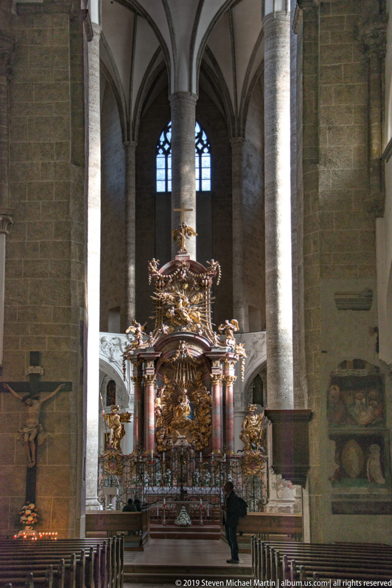 Darkened nave with massive stone columns framing the illuminated high altar, a crucifix at left, and a visitor near center.