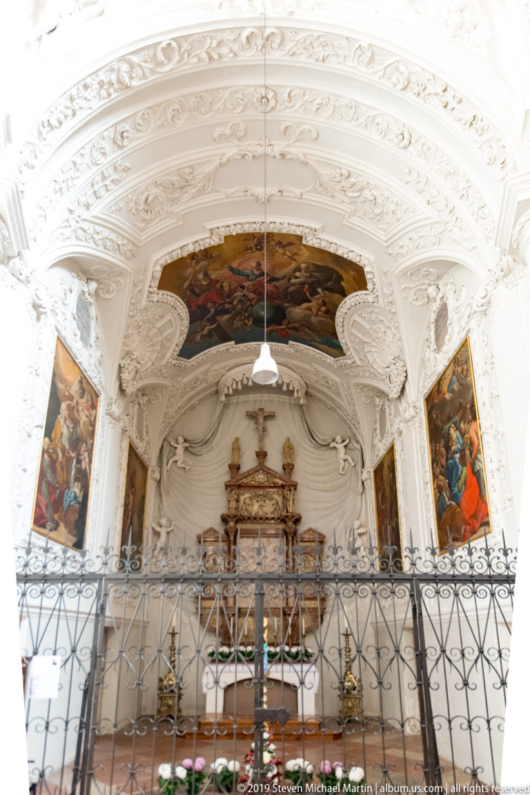 Pale side chapel with restrained altar, large ceiling painting, two vertical side paintings, hanging lamp, and iron screen.