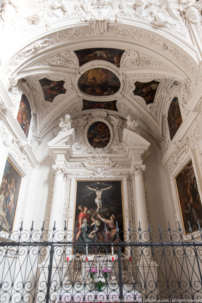 White stucco side chapel with a large Crucifixion painting, smaller ceiling paintings, sculpted pediment, and iron screen.
